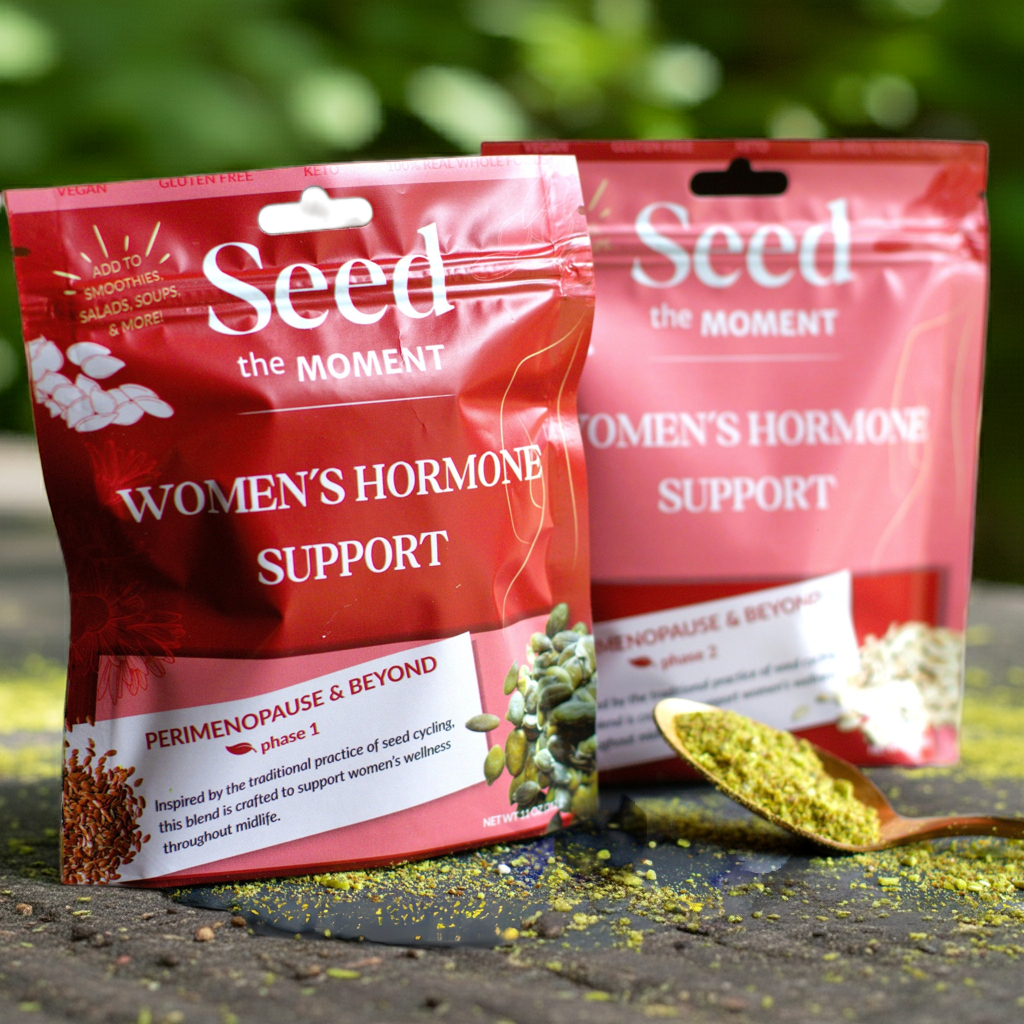 Two red packages labeled 'Seed the Moment Women's Hormone Support' on a dark surface with greenery in the background.