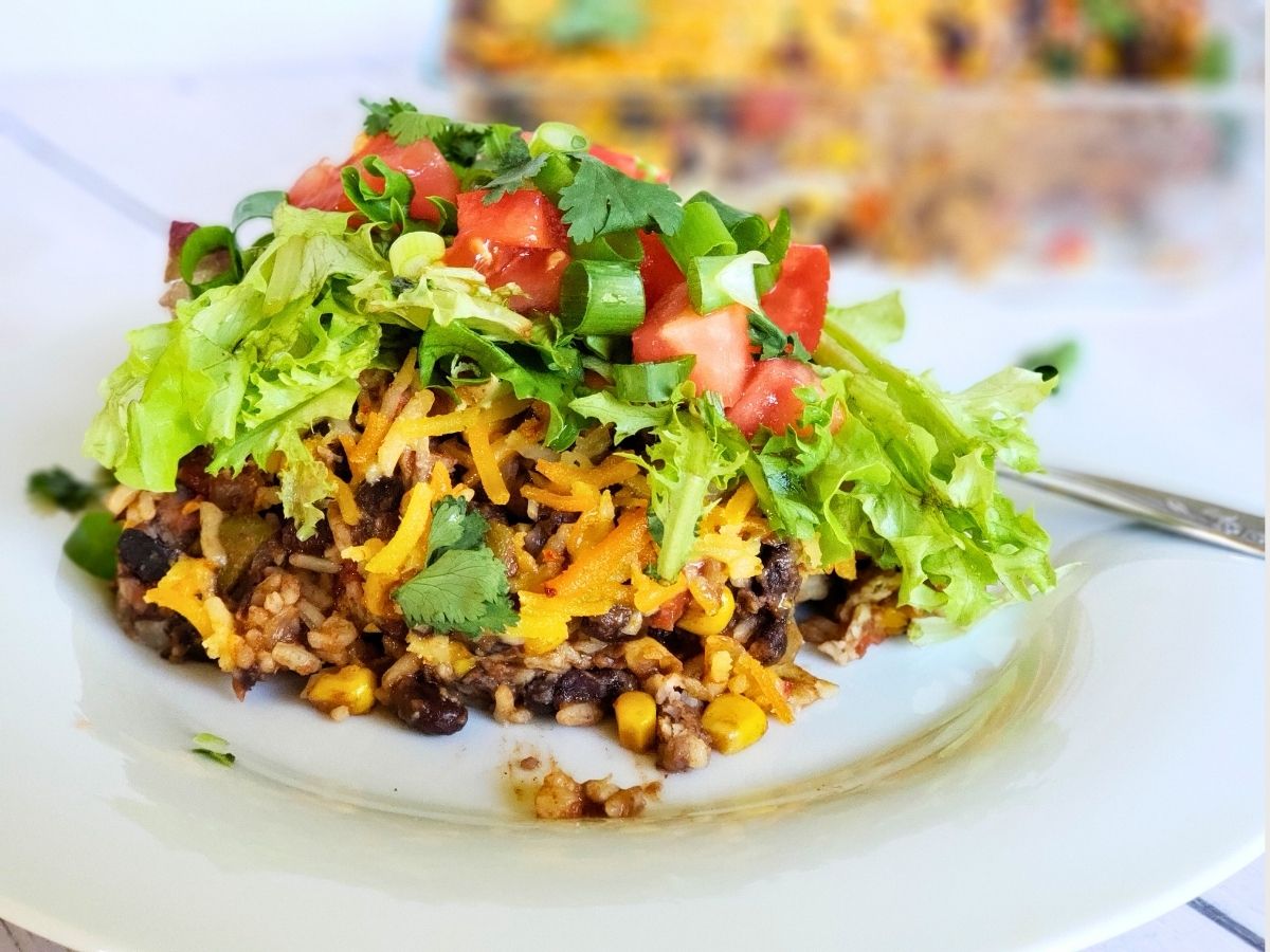 Taco casserole with rice, beans, dairy free cheese, lettuce, and tomatoes on a white plate.