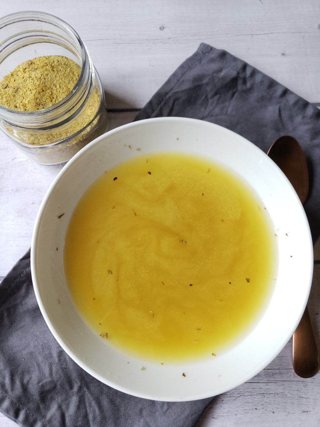 White bowl with vegan chicken broth on a gray surface, next to a jar of vegan bouillon powder and a wooden spoon.